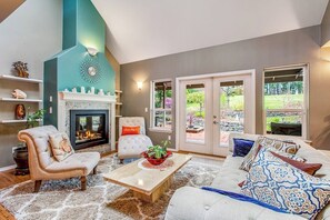 A living room with a fireplace, a sofa, a coffee table, and a view of the outdoors through large windows. - 3BR Golf Course View (Allyn)