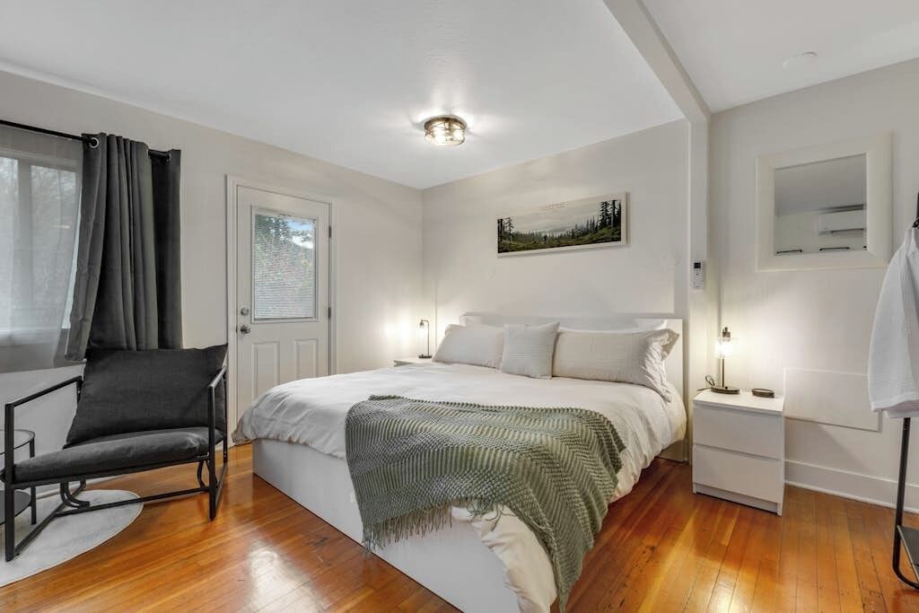 Une chambre à coucher avec un lit, des tables de chevet, une chaise, une fenêtre avec des rideaux et un tableau au mur.