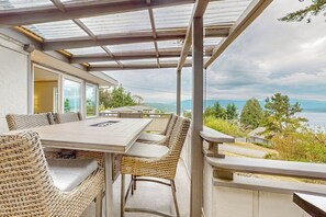 An outdoor patio with a table and wicker chairs, overlooking a scenic landscape. - Modern sound view home with expansive deck, mtn view (Port Townsend)