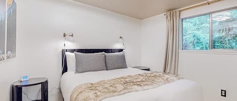 A bedroom with a bed, bedside tables, a window with curtains, and a wall-mounted light fixture.