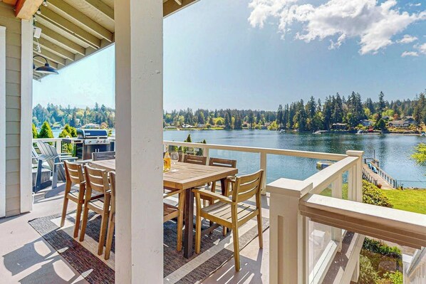 A covered outdoor area with a wooden table and chairs, overlooking a lake with trees and houses in the background. - Waterfront Home Nearby Island Paradise (Gig Harbor)