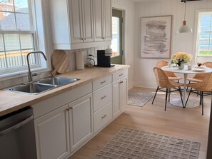 A kitchen with white cabinetry, a stainless steel sink, a coffee maker, and a dining area with wicker chairs and a white table. - Maple & Moss | Cozy 2-Bedroom Retreat in Moncton (Moncton)