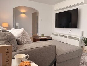 A modern living room with a grey sofa, a coffee table, a flat-screen TV mounted on the wall, and a lamp. - Maple & Moss | Cozy 2-Bedroom Retreat in Moncton (Moncton)