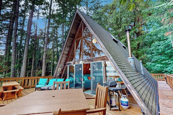 Una cabaña con techo inclinado, una terraza con asientos y una zona de barbacoa.