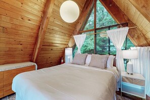 A bedroom with a large bed, a bench, a bedside table, a lamp, and a window with curtains.