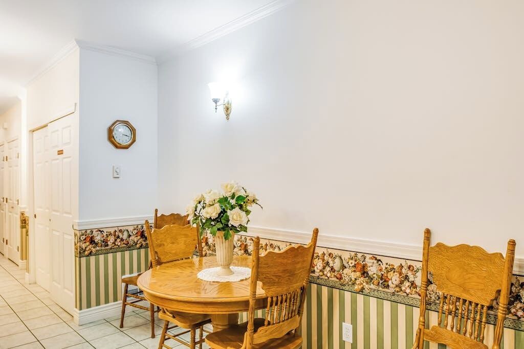 Uma sala de jantar com uma mesa redonda de madeira, quatro cadeiras, um vaso com flores e um relógio de parede.
