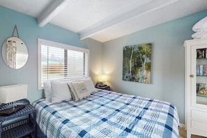 Un dormitorio con cama, mesita de noche, lámpara, espejo, ventana con persianas, un cuadro y estantería.