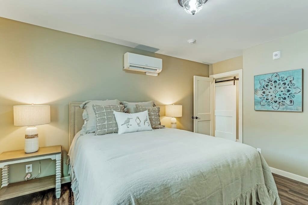 A bedroom with a bed, bedside tables, a wall-mounted air conditioner, a door, and a wall art piece.