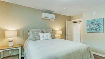 A bedroom with a bed, bedside tables, a wall-mounted air conditioner, a door, and a wall art piece.