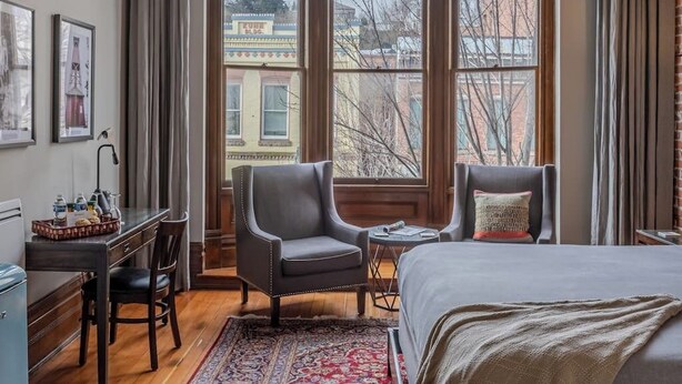 Ein Hotelzimmer mit großem Fenster, einem Bett, einem Schreibtisch, zwei Stühlen und einem kleinen Tisch.