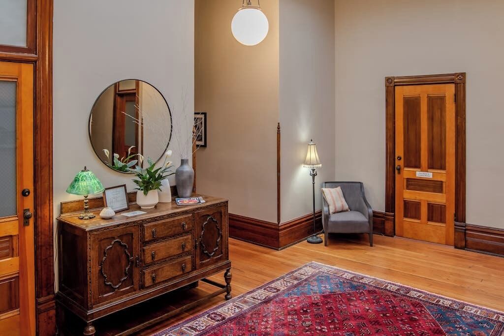A room with a wooden cabinet, a round mirror, a lamp, a chair, and a door.