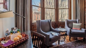 Ein Hotelzimmer mit zwei Ledersesseln, einem kleinen Tisch, einem Fenster mit Blick auf Gebäude und einer Ablage mit Snacks und einer Wasserflasche.