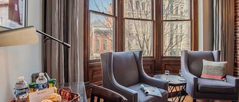 A hotel room with two leather armchairs, a small table, a window with a view of buildings, and a tray with snacks and a water bottle.