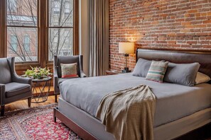 Une chambre d’hôtel avec un mur en briques, un grand lit avec de nombreux oreillers, une table de chevet avec une lampe, une chaise et une vue sur l’extérieur grâce à de grandes fenêtres.