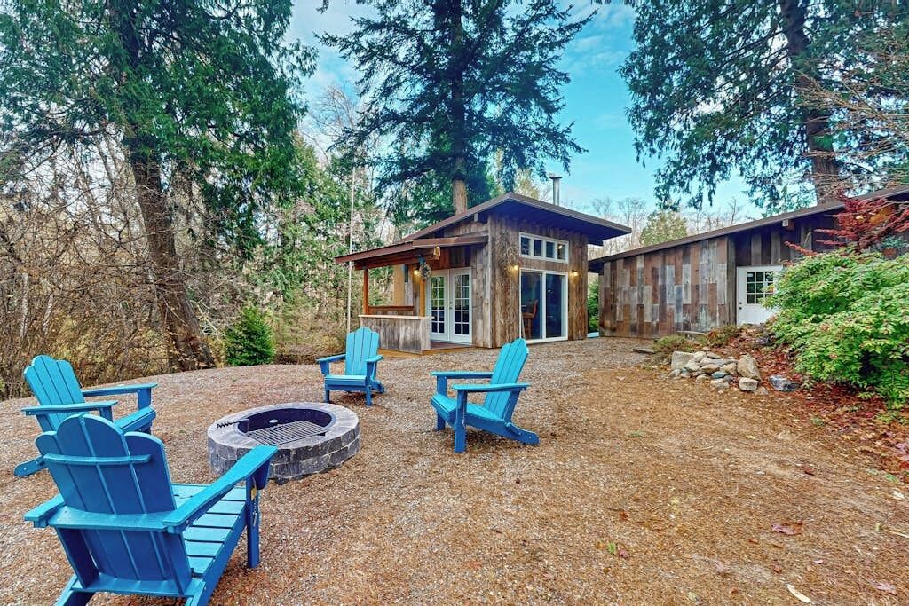 Una cabaña con fogata y sillas Adirondack azules en un entorno boscoso.