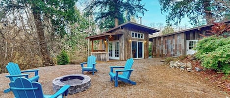 Una cabaña con fogata y sillas Adirondack azules en un entorno boscoso.