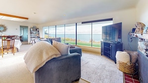 A modern living room with a large window, a flat-screen TV, and a cozy seating area. - 4BR Home with bay and mountain views, beach access (Sequim)