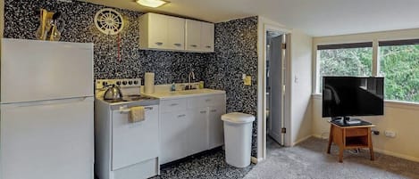 A compact kitchen with white cabinets, a stove, and a sink. There's a trash can and a television on a wooden stand.