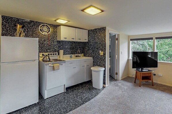 A compact kitchen with white cabinets, a stove, and a sink. There's a trash can and a television on a wooden stand.