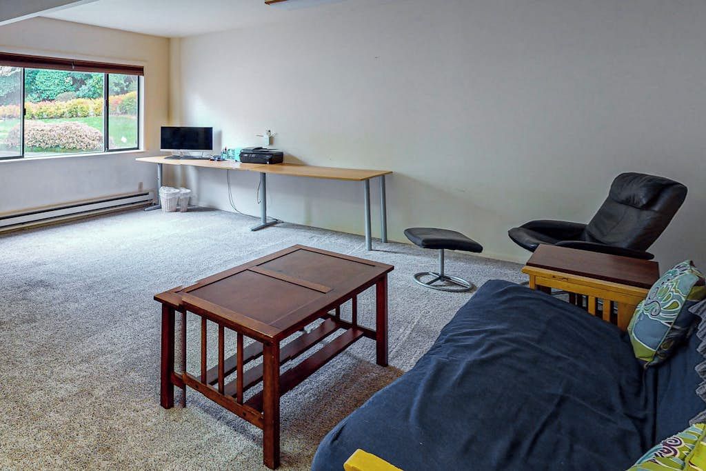 A room with a sofa, a small wooden table, a desk with a computer and a chair, and a window with a view of greenery.