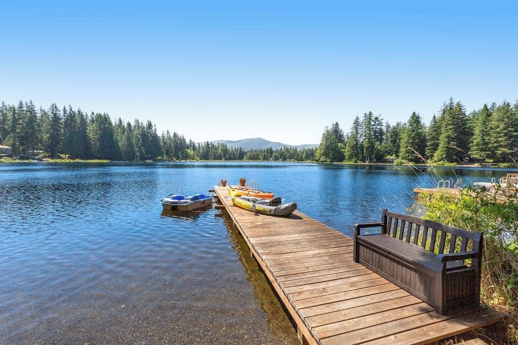 A wooden dock extending into a calm lake with boats docked, surrounded by dense forest and a clear sky.