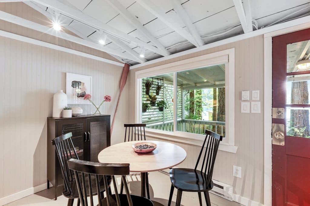 A dining area with a round table, four chairs, a cabinet, and a window with a view of greenery.