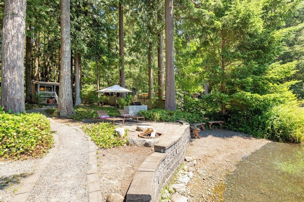 A forested area with a stone fire pit, a picnic table, and a small deck with a chair.