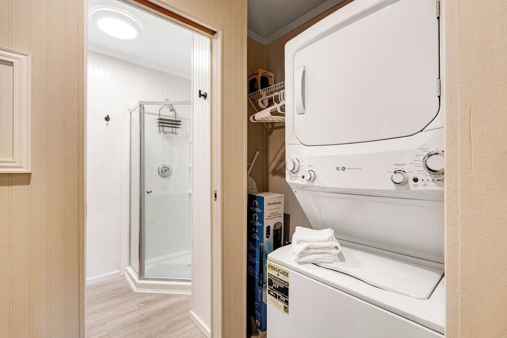 A laundry room with a front-loading washer and dryer, a glass shower enclosure, and a storage area with shelves.