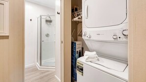 Een wasserette met een voorlader wasmachine en droger, een glazen douchecabine en een bergruimte met planken.