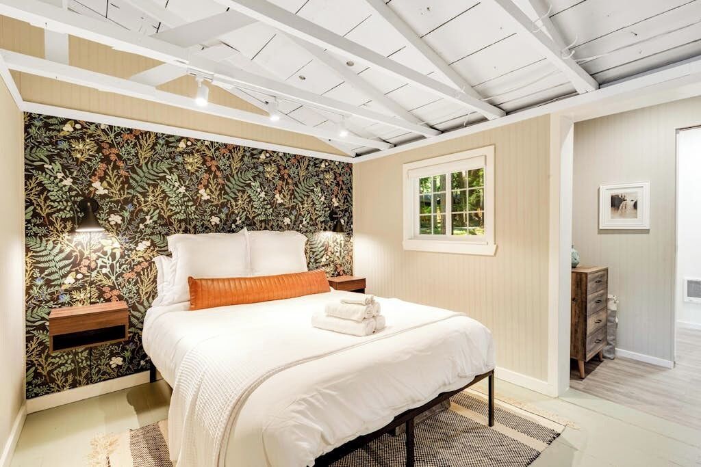 A bedroom with a large bed, a floral-patterned headboard, bedside lamps, a window, and a wooden dresser.