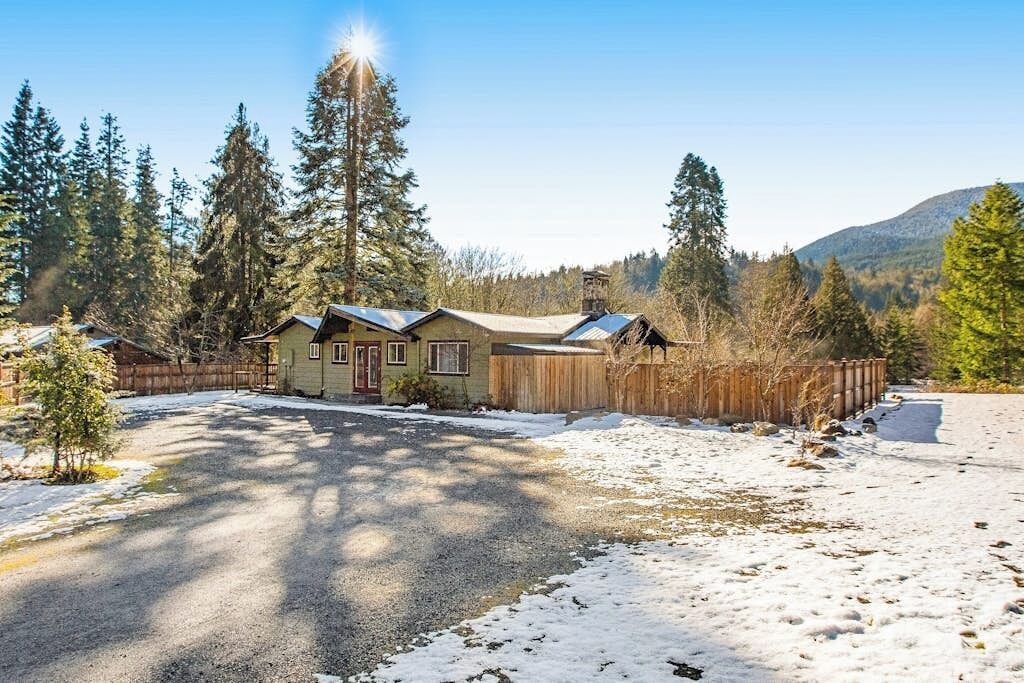 Une maison de plain-pied, dotée d’un toit en pente, entourée d’une clôture en bois, située dans un paysage enneigé, avec des arbres à feuilles persistantes et une montagne en arrière-plan.