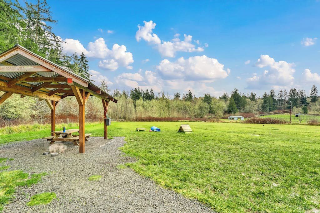 Un espace pique-nique abrité, équipé d’une table et de bancs, entouré d’un champ herbeux et d’arbres.