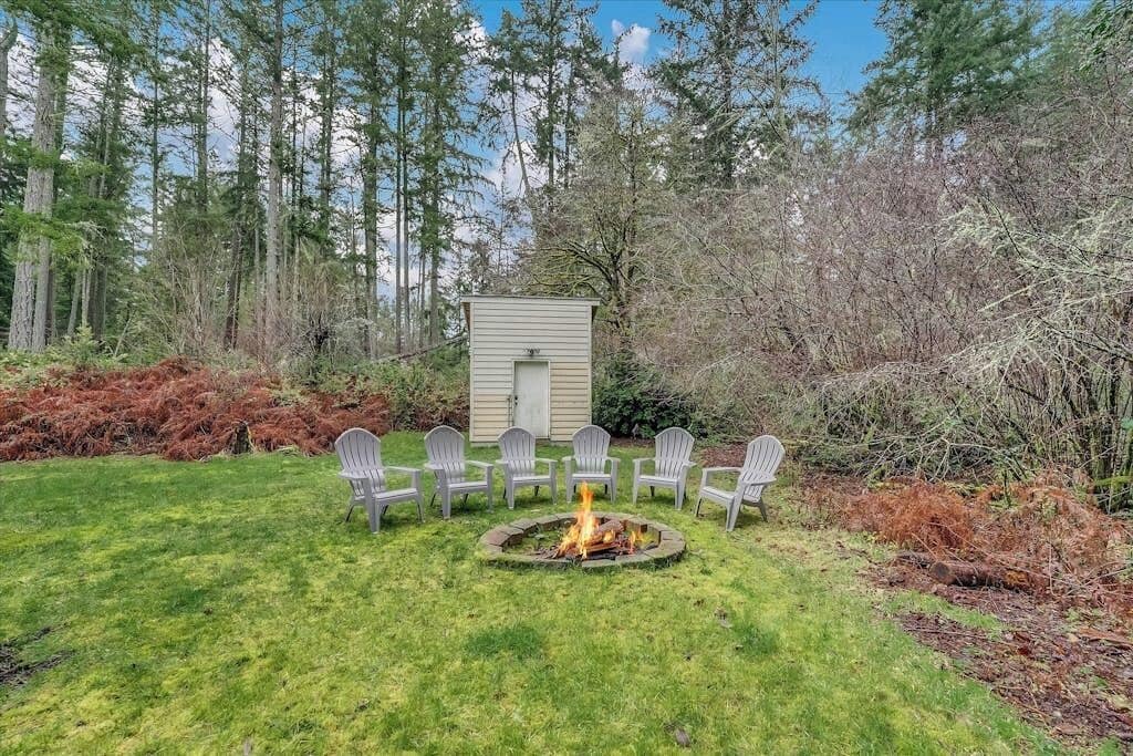 Un jardin avec un foyer et des chaises disposées autour, le tout entouré d’une zone boisée.