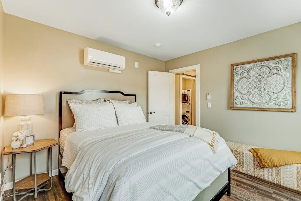Un dormitorio con cama, mesita de noche, lámpara, equipo de aire acondicionado y una puerta que da a un baño.