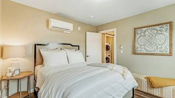 A bedroom with a bed, bedside table, lamp, air conditioning unit, and a door leading to a bathroom.