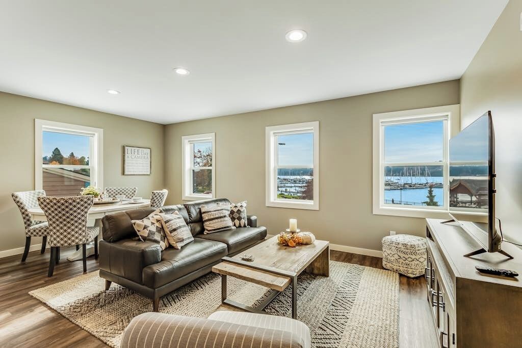 A living room with a sofa, coffee table, and ottoman, a dining area with chairs and a table, and large windows with a view of a marina.
