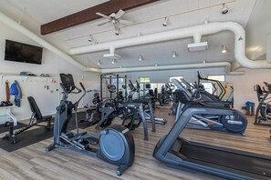 Academia com diversos equipamentos de ginástica, um ventilador de teto e uma TV na parede.
