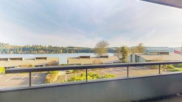 Une vue depuis une fenêtre donnant sur un parking et des immeubles, avec une étendue d’eau et des arbres visibles au loin.