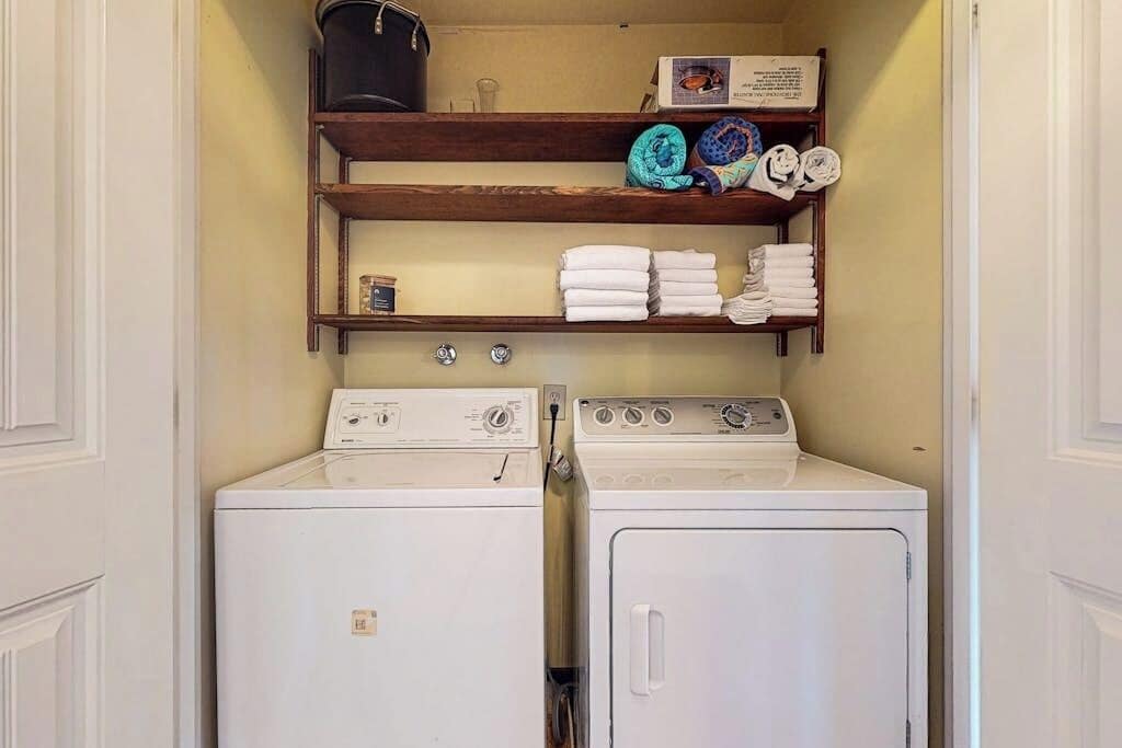 A laundry room with two white washing machines, a shelf with folded towels, and a box of laundry detergent.