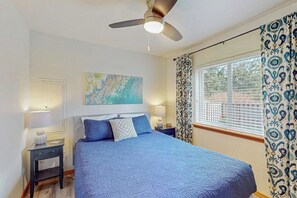 Ein Schlafzimmer mit einem Bett, einem Deckenventilator, einem Fenster mit gemusterten Vorhängen und einem an der Wand hängenden Kunstwerk.