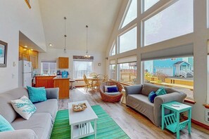Ein großzügiger Wohnbereich mit einer Couch, einem Couchtisch und einem Korbstuhl. Es gibt eine Küchenzeile mit einem Kühlschrank und einem Esstisch. Das Zimmer verfügt über große Fenster mit Blick ins Freie.