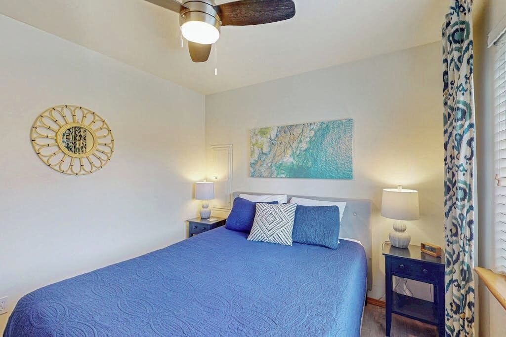 A bedroom with a bed, a ceiling fan, a wall art piece, and a nightstand with a lamp.