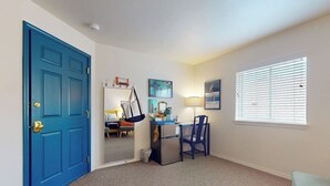 A room with a blue door, a desk with a chair, a small refrigerator, and a window with blinds.