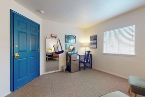 Una habitación con puerta azul, un escritorio con silla, un refrigerador pequeño y una ventana con persianas.
