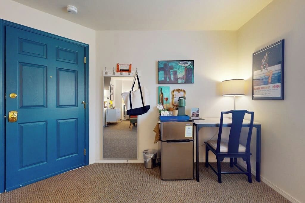 A hotel room with a blue door, a desk with a chair, a small refrigerator, a trash can, and a mirror reflecting the room.