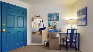 A hotel room with a blue door, a desk with a chair, a small refrigerator, a trash can, and a mirror reflecting the room.