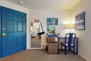 Habitación de hotel con puerta azul, escritorio con silla, refrigerador pequeño, basurero y espejo que refleja la habitación.