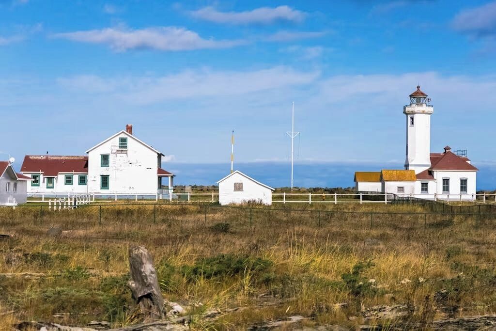 Un faro costero con fachada blanca y techo rojo, rodeado de terreno cubierto de hierba y un edificio blanco con ventanas verdes.