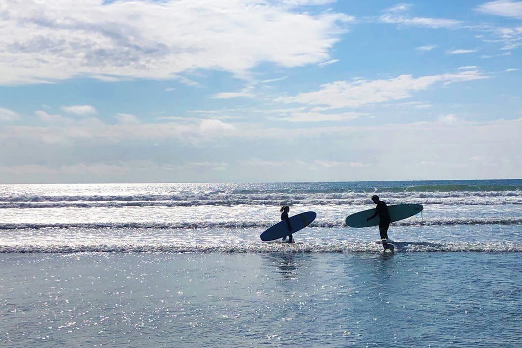 Two individuals walking on a beach with surfboards, ocean waves in the background, and a clear sky.