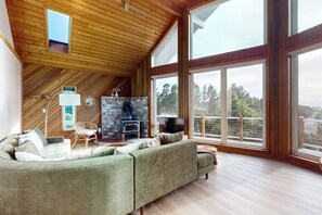 A spacious living room with a large green sofa, a stone fireplace, and large windows offering a view of the outdoors. - 1BR  with loft retreat with wrap around balcony & Washer Dryer (Westport)